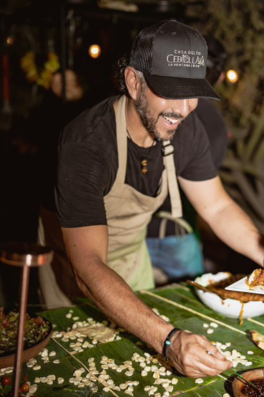 Chef Raymond smiling behind roasted meats and vegetables hanging over the open fire at Cebollajo